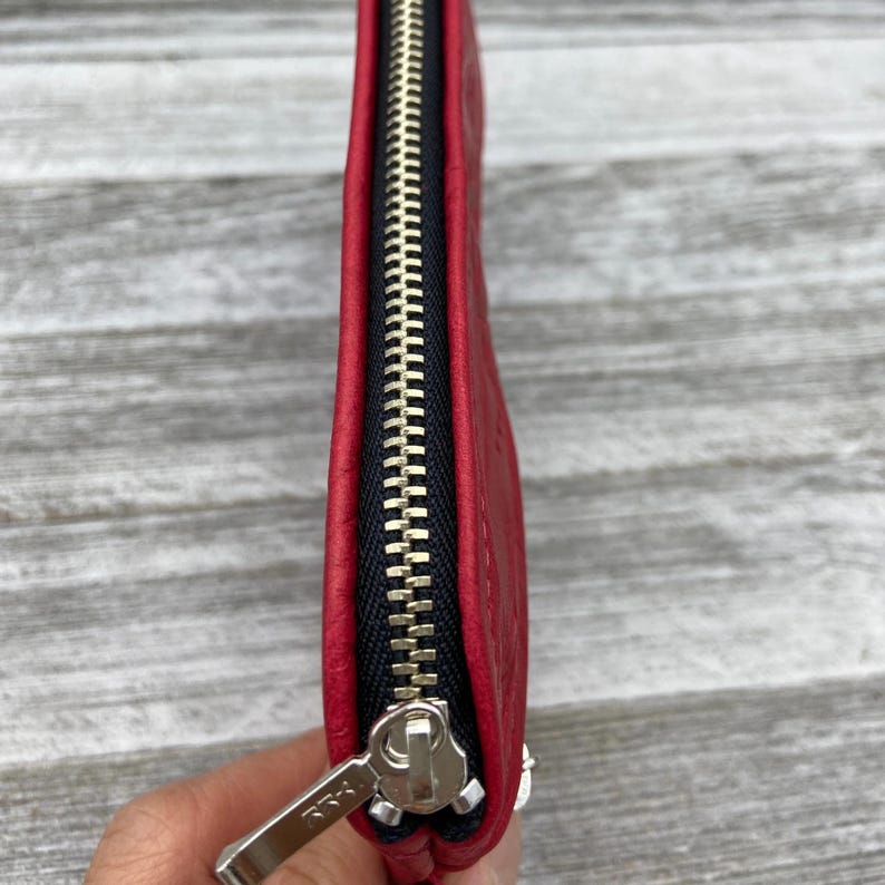 May include: A close-up of a red leather wallet with a black zipper and silver zipper pull. The wallet is rectangular and appears to be made of high-quality leather. The zipper is closed, and the wallet is in good condition.
