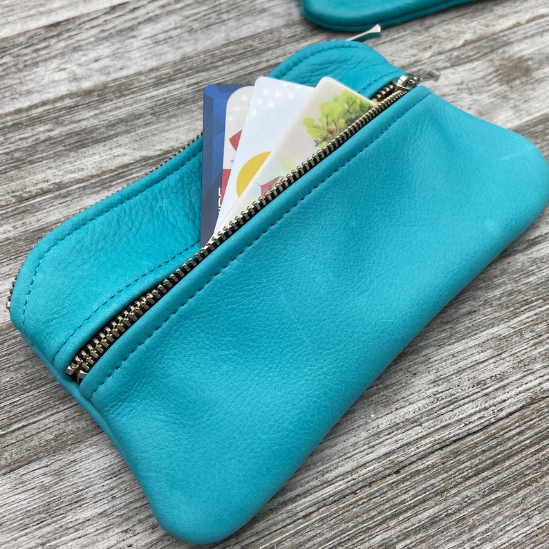 May include: A turquoise leather pouch with a silver zipper, partially open to reveal several cards. The pouch is made of a soft, textured leather. The cards have various designs and colors. The pouch is designed to hold cards and small items.