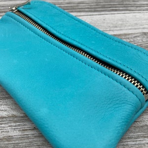 May include: A turquoise leather pouch with a silver zipper closure. The pouch is rectangular and has a slightly textured surface. The zipper runs along the top edge of the pouch.