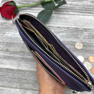 May include: A purple leather wallet with a gold zipper is open, revealing cash and a credit card. The wallet is held in a hand, with a red rose and coins in the background. The wallet is a long, rectangular shape.