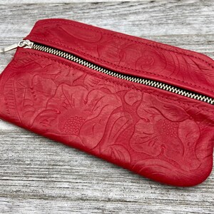 May include: A red leather clutch with a floral embossed design. The clutch has a silver zipper closure and a rectangular shape. The leather is a vibrant red color, and the floral pattern is detailed and textured.