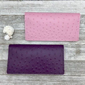 May include: Two leather wallets, one light pink and one deep purple, are displayed on a weathered wooden surface. The wallets have a textured, dotted pattern and a rectangular shape with a flap closure.
