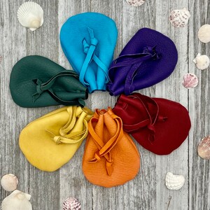 May include: Seven small leather pouches in a rainbow of colors, arranged in a circle on a wooden surface. The pouches are tied with leather cords and have a drawstring closure.