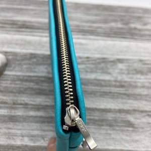 May include: A close-up of a turquoise clutch with a silver zipper. The zipper pull has a star design. The clutch is made of a smooth material and has a black interior.
