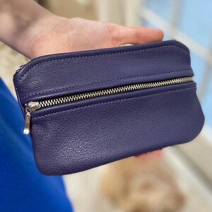 May include: A deep purple leather pouch with a silver zipper closure. The pouch is rectangular with rounded edges and a textured surface. The zipper pull is silver. The pouch is held in a hand.