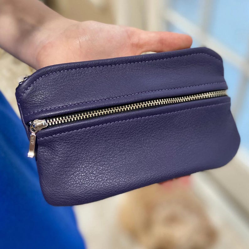 May include: A deep purple leather pouch with a silver zipper closure. The pouch is rectangular with rounded edges and a textured surface. The zipper pull is silver. The pouch is held in a hand.