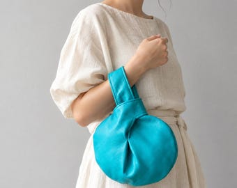 Könnte beinhalten: Eine leuchtend türkisfarbene Handgelenktasche mit abgerundeter, plissierter Silhouette und einem einzelnen integrierten Griff. Die Tasche hat eine weiche, strukturierte Oberfläche und ein gerafftes Design, das eine runde Form bildet.