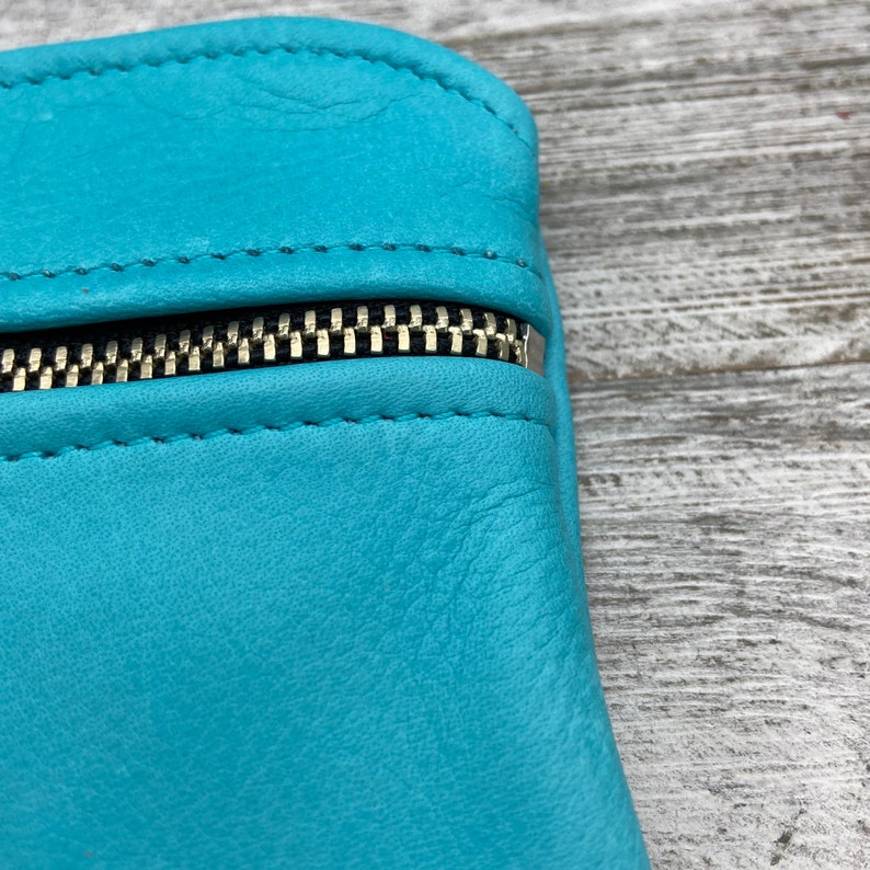 May include: Close-up of a turquoise leather pouch with a gold zipper. The leather has a textured appearance and is stitched along the edges. The zipper is open, revealing the interior of the pouch.