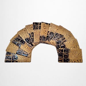 May include: A decorative arch made of textured, tan-colored material with dark brown accents. The arch is composed of multiple layered pieces, creating a textured, layered effect. The design resembles a reptile skin pattern.