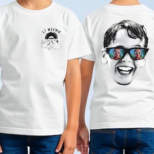 May include: White t-shirt featuring a graphic on the front with the text "L7 Weenie" and a hand gesture. The back of the shirt displays a black and white image of a smiling boy wearing sunglasses with a mirrored reflection. The shirt is worn by a person.