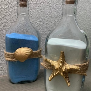May include: Two glass bottles with cork stoppers filled with blue and white sand. The bottles are decorated with twine and a gold seashell and a gold starfish.