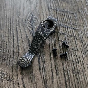 Puede incluir: Un cuchillo de bolsillo de titanio negro con un patrón dorado. El cuchillo está sobre una superficie de madera con tres pequeños tornillos negros.