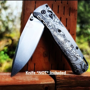 Puede incluir: Un cuchillo plegable negro con un patrón grabado decorativo en el mango. El cuchillo está cerrado y descansa sobre una superficie de madera. Cuchillo *NO* Incluido.