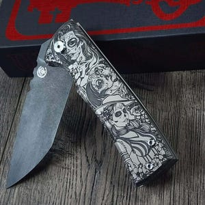 May include: A folding pocket knife with a detailed Day of the Dead design. The handle features a black and white illustration of a woman with sugar skull makeup. The blade is open, revealing a gray, matte finish.