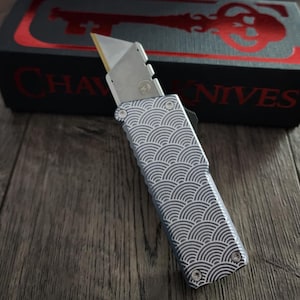 May include: A silver utility knife with a wave pattern on the handle, resting on a wooden surface. The knife has a gold blade and is partially extended. The box behind the knife has the words "CHAVES KNIVES" in red.
