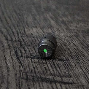 May include: A small, cylindrical black object with a bright green circular light on one end. The object is resting on a dark, textured wooden surface. The green light is the focal point, suggesting a technological or optical component.