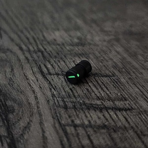 Può includere: Un piccolo oggetto cilindrico nero con una luce verde sul lato. L'oggetto è appoggiato su una superficie di legno scuro.