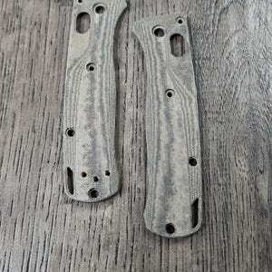 May include: Two gray knife handle scales with a wood grain pattern. Each scale has multiple holes for screws and a curved shape. The scales are made of a durable material and are designed for a knife.