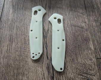 May include: Two pale green knife handle scales with multiple holes for attachment. The scales are lying on a wooden surface. A blue box with the word "BENCHMADE" is in the background.