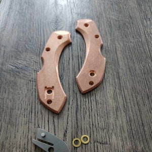 May include: Copper-colored knife handle scales, a silver-colored knife lock, and two small gold-colored washers are arranged on a wooden surface. The handle scales have multiple holes and a curved design.