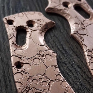 Puede incluir: Dos escamas de metal de color cobre con un patrón de calavera. Las escamas están diseñadas para un mango de cuchillo.