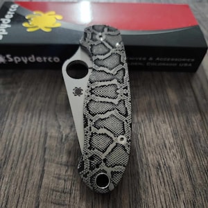 May include: A black and grey Spyderco knife with a snakeskin pattern on the handle. The knife is closed and the handle is visible. The knife is on a wooden surface.