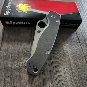 May include: A gray Spyderco pocket knife with a black handle and a Spyderco logo on the blade. The knife is closed and sitting on a wooden surface. The knife is next to a red and black box with the Spyderco logo and the text "& Accessories Colorado USA".
