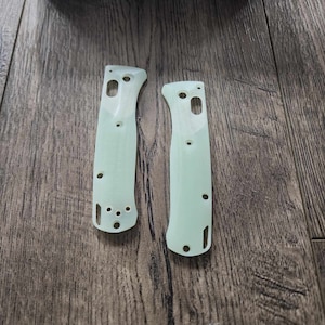 May include: Two light green knife handle scales with multiple holes and a curved design. The scales are made of a translucent material and are positioned on a wooden surface. A blue box with the word "BENCHMADE" is in the background.