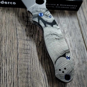 May include: A silver pocket knife with a black and gray patterned handle. The knife has a blue screw and the words "S30V CPM S11EV" are engraved on the handle. The knife is sitting on a wooden surface.