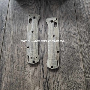 May include: Two textured knife handle scales with multiple holes and a cutout, in a light gray color. The text "DNALASERNG.COM" is printed on the scales. A blue box with the word "BENCHMADE" is in the background.