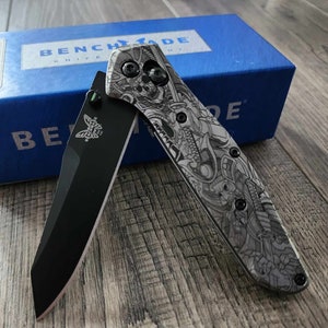 May include: A black and gray pocket knife with a detailed, intricate design on the handle. The knife is open and the blade is visible. The knife is sitting on a wooden surface. The knife is a Benchmade brand.