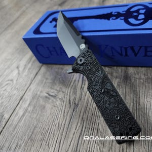 May include: A black pocket knife with an intricate laser-etched design on the handle. The knife is closed and resting on a wooden surface. The knife is packaged in a blue box with the text "CHA KNIVES" printed on it.