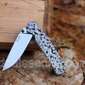 May include: A silver pocket knife with a black and white floral pattern on the handle. The knife is open and the blade is visible. The brand name "RUWKE" is printed on the blade.
