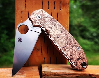 May include: A folding pocket knife with a silver-toned blade featuring a circular thumb hole and a spider logo. The handle is made of copper with an intricate laser-engraved design of an octopus and the text "NALASERING.COM" visible on the side.