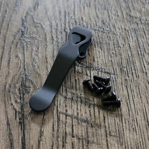 以下が含まれることがあります： 黒いポケットクリップと小さな黒いネジのセットが木製の表面に配置されています。 クリップは、片側にフック、もう一方に丸みを帯びた先端が付いた、湾曲した細長い形状をしています。