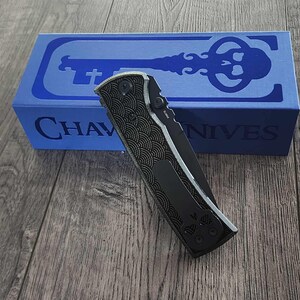 May include: A black folding knife with a patterned handle and a silver blade, resting on a wooden surface. A blue box with the text "CHAVES KNIVES" and a key design is in the background.