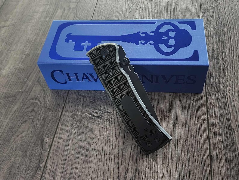 May include: A black folding knife with a patterned handle and a silver blade, resting on a wooden surface. A blue box with the text "CHAVES KNIVES" and a key design is in the background.