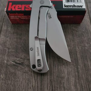 Puede incluir: Una navaja plateada con una hoja parcialmente dentada, con un clip para el bolsillo y la marca "Kershaw". El cuchillo se muestra sobre una superficie de madera, con una caja roja y negra en el fondo.