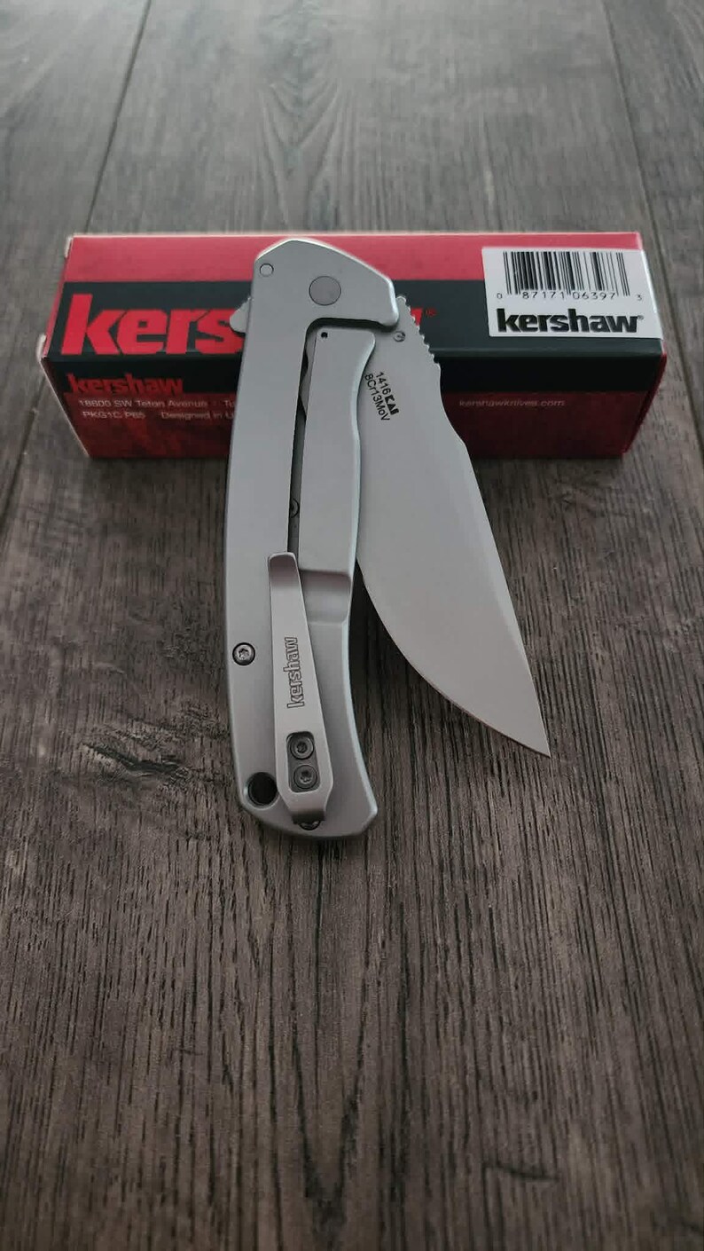 Puede incluir: Una navaja plateada con una hoja parcialmente dentada, con un clip para el bolsillo y la marca "Kershaw". El cuchillo se muestra sobre una superficie de madera, con una caja roja y negra en el fondo.