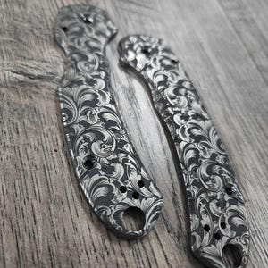 May include: Two decorative metal knife handles with an ornate floral pattern. The handles are silver and black, with a detailed scrollwork design. Each handle has a hole at the base and several smaller holes along the body.