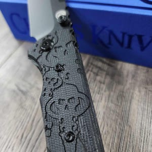 May include: A black pocket knife with a skull and crossbones pattern on the handle. The knife is partially resting on a blue box with the word "CKNIVES" printed on it.