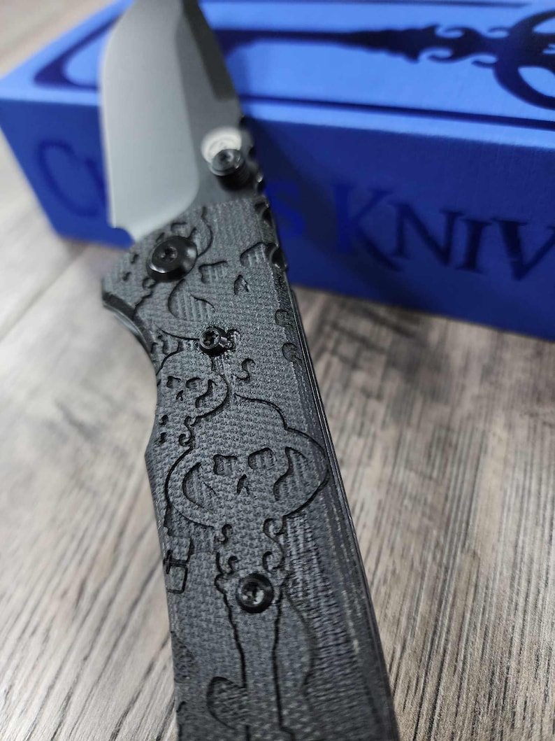 May include: A black pocket knife with a skull and crossbones pattern on the handle. The knife is partially resting on a blue box with the word "CKNIVES" printed on it.