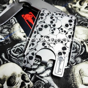 May include: A silver folding knife and a silver card holder with a skull pattern. The card holder has a key design and is on a black and white skull and rose patterned background.