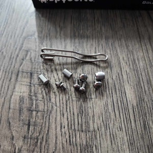 May include: A set of silver-colored screws and a silver-colored wire loop, likely for assembling a pocket knife. The screws are on a wooden surface. The wire loop is bent into a U-shape.