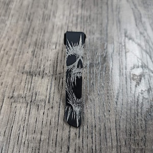 Puede incluir: Un abridor de botellas de metal negro con un diseño detallado de calavera blanca. La calavera presenta dientes afilados y una corona puntiaguda. El abridor es rectangular con una parte superior curva.