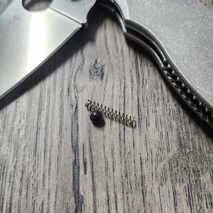May include: A close-up of a small, silver spring and a black ball bearing. The spring is lying on a light brown wooden surface.