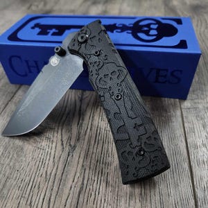 May include: A black pocket knife with a textured handle and a partially open blade. The knife is sitting on a wooden surface next to a blue box with the word "CHIVES" printed on it.