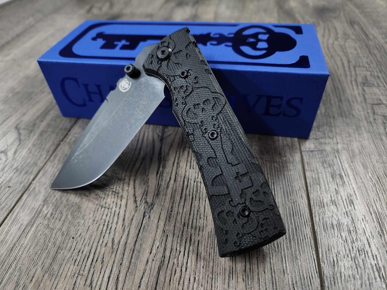May include: A black pocket knife with a textured handle and a partially open blade. The knife is sitting on a wooden surface next to a blue box with the word "CHIVES" printed on it.