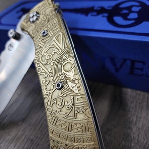 Puede incluir: Un cuchillo de bolsillo dorado con un diseño grabado intrincado que presenta una calavera y patrones florales. El cuchillo está cerrado y descansa sobre una caja azul con la palabra "IVES" impresa en ella.