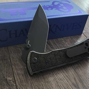May include: A black folding knife with a partially open blade, resting on a wooden surface. The handle features a textured pattern and a pocket clip. A blue box with the text "CHAVES KNIVES" is in the background.